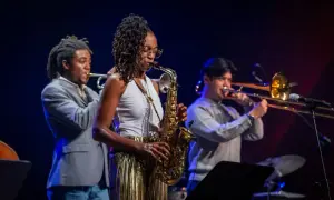 Zespół jazzowy gra na scenie. Kobieta gra na saksofonie, mężczyzna na trąbce, a drugi na puzonie. Inspiracja do tego, jak napisać tekst piosenki.