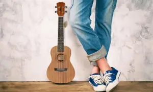 Drewniane ukulele obok osoby w jeansach i niebieskich trampkach. Idealne do grania letnich hitów na ukulele.