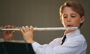Chłopiec w białej koszuli i krawacie gra na srebrnej **fletni**.