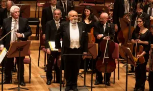 Orkiestra przygotowuje się do wykonania Dies Irae. Dyrygent w białej kamizelce i fraku stoi na podwyższeniu, otoczony muzykami z instrumentami smyczkowymi.