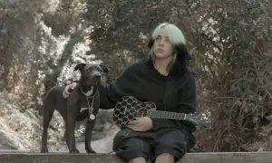 Billie Eilish z pieskiem i ukulele w rękach, siedząca na ławce w otoczeniu natury.