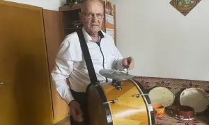 Starszy mężczyzna w białej koszuli gra na dużym, drewnianym bębnie. Obok niego stoją mniejsze instrumenty perkusyjne.