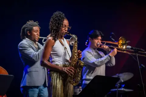 Zespół jazzowy gra na scenie. Kobieta gra na saksofonie, mężczyzna na trąbce, a drugi na puzonie. Inspiracja do tego, jak napisać tekst piosenki.