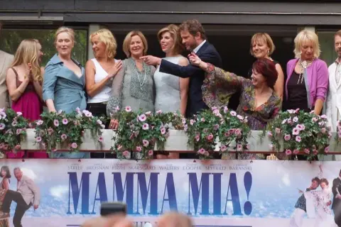 Obsada filmu "Mamma Mia!" pozdrawia fanów. W tle plakat z bohaterami, przypominający o wesołych piosenkach z filmu.
