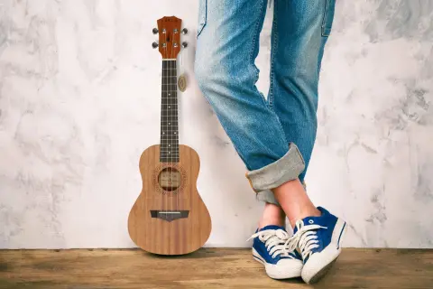 Drewniane ukulele obok osoby w jeansach i niebieskich trampkach. Idealne do grania letnich hitów na ukulele.