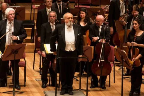 Orkiestra przygotowuje się do wykonania Dies Irae. Dyrygent w białej kamizelce i fraku stoi na podwyższeniu, otoczony muzykami z instrumentami smyczkowymi.