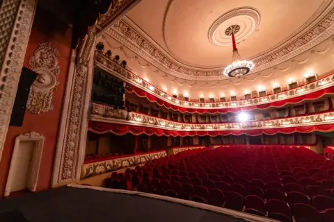 Wnętrze teatru z czerwonymi fotelami i bogato zdobionymi lożami. Czekamy na rozpoczęcie spektaklu, by usłyszeć piękną **arię**.
