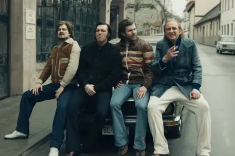 Czterech mężczyzn w stylu lat 70. siedzi na samochodzie. Jeden z nich pali papierosa, przypominając scenę z filmu "Jak pokochałam gangstera".