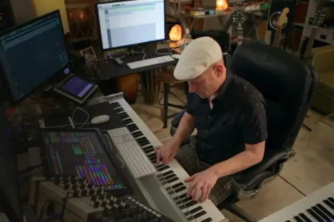 Muzyk w białej czapce tworzy muzykę w studiu. Komputery, klawiatury i miksery sugerują, że zastanawia się, dlaczego nie soundtrack.