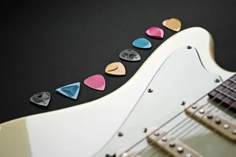 Kolorowe kostki do gitary ułożone wzdłuż białego korpusu instrumentu. Idealne akcesoria, by zacząć naukę gry na gitarze.