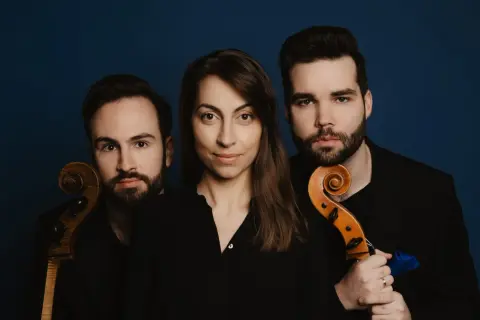 Trzech muzyków, tworzących triolę, pozuje z instrumentami smyczkowymi na tle granatowym.