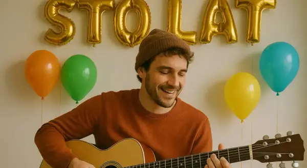 Uśmiechnięty mężczyzna w czapce gra na gitarze na tle złotych balonów z napisem "STO LAT" i kolorowych balonów.