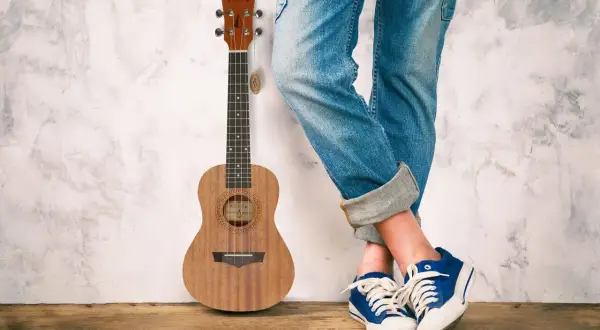 Drewniane ukulele obok osoby w jeansach i niebieskich trampkach. Idealne do grania letnich hitów na ukulele.