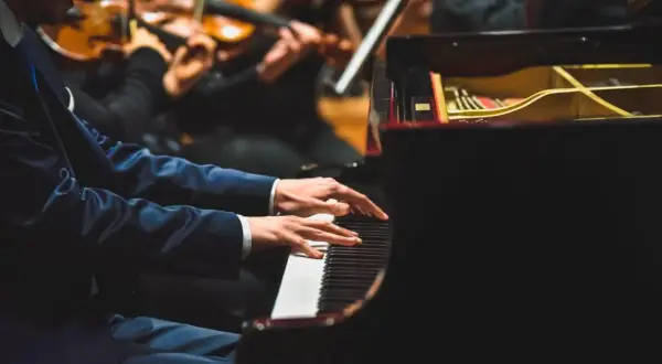 Pianista w eleganckim garniturze gra na fortepianie, a przed nim leżą nuty do nauki gry na pianinie. W tle orkiestra smyczkowa.