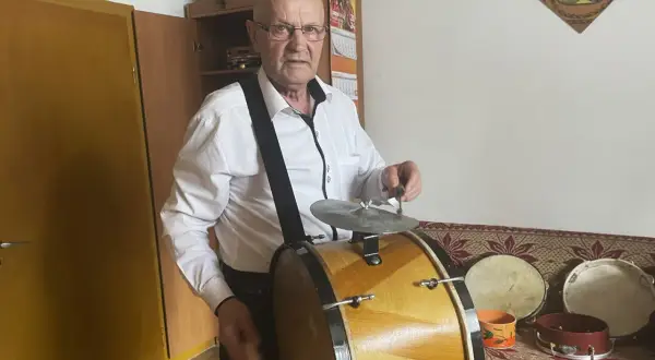 Starszy mężczyzna w białej koszuli gra na dużym, drewnianym bębnie. Obok niego stoją mniejsze instrumenty perkusyjne.