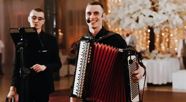 Uśmiechnięty muzyk gra na akordeonie, tworząc radosną muzykę biesiadną. Obok stoi kolega z tamburynem.