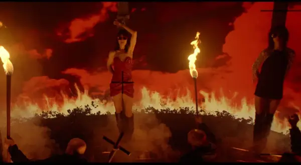 Scena z płonącym tłem, gdzie kobieta w czerwonej sukience jest przywiązana do krzyża. Wokół płoną pochodnie, a w oddali widać drugą kobietę. To obraz **lux aeterna**.
