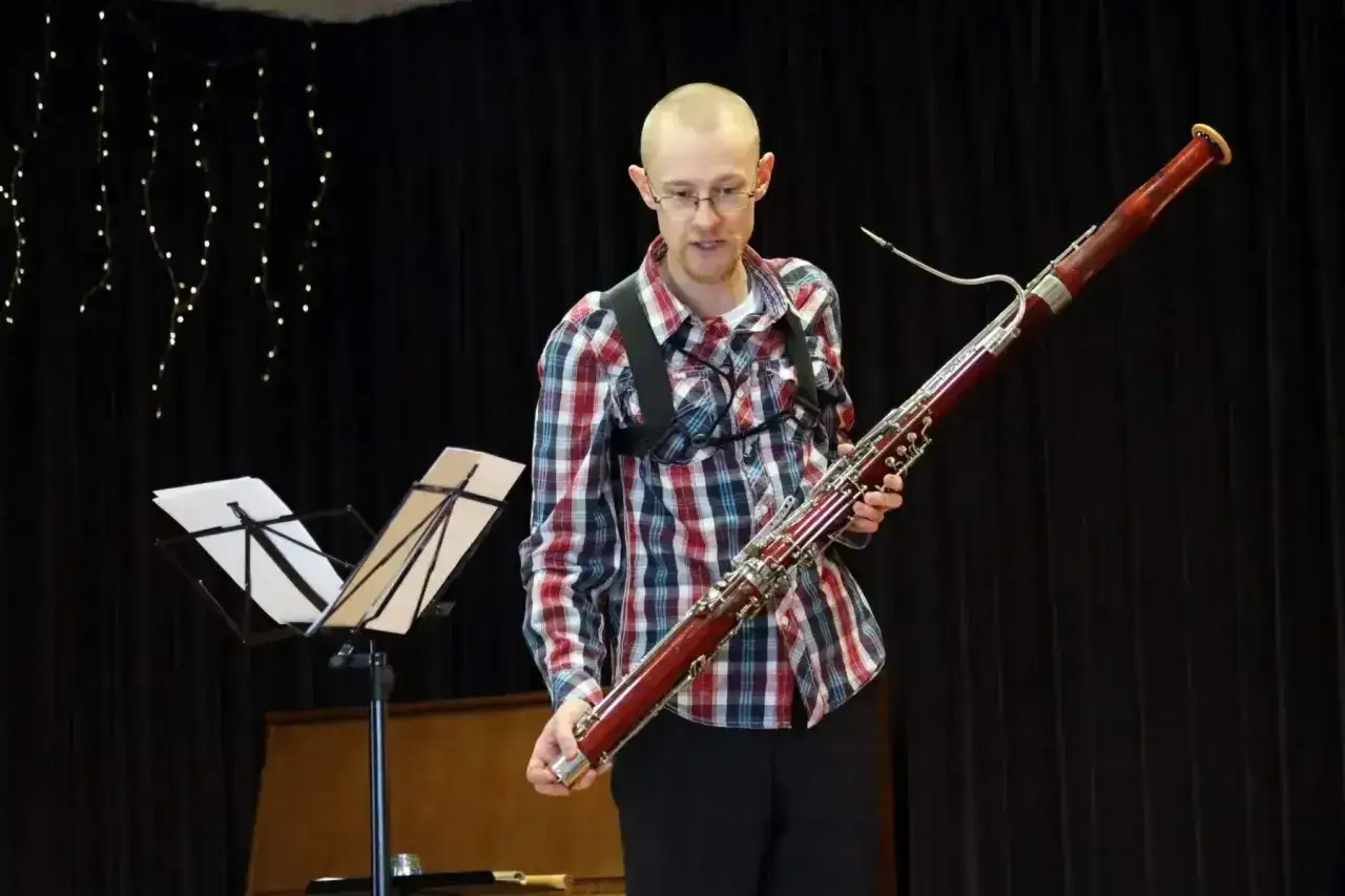 Mężczyzna w kratę trzyma fagot, instrument muzyczny. Na scenie stoją nuty i świecące lampki.