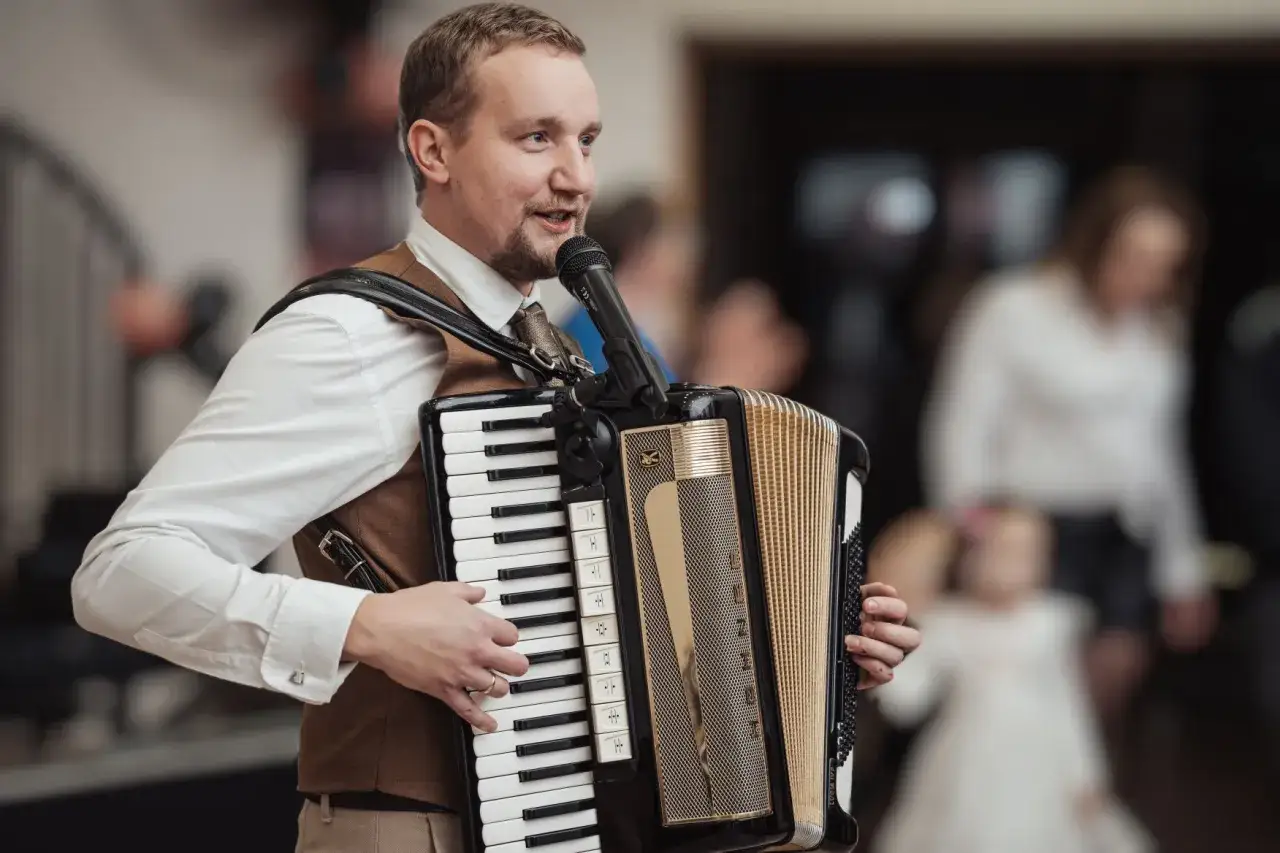 Akordeonista grający piosenki na akordeon biesiadne, z mikrofonem i w eleganckim stroju. W tle widać bawiących się gości.