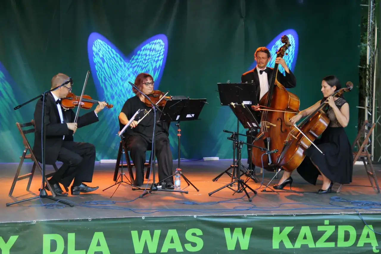 Kwartet smyczkowy gra na scenie. Muzycy w eleganckich strojach wykonują utwór, tworząc harmonijną całość.