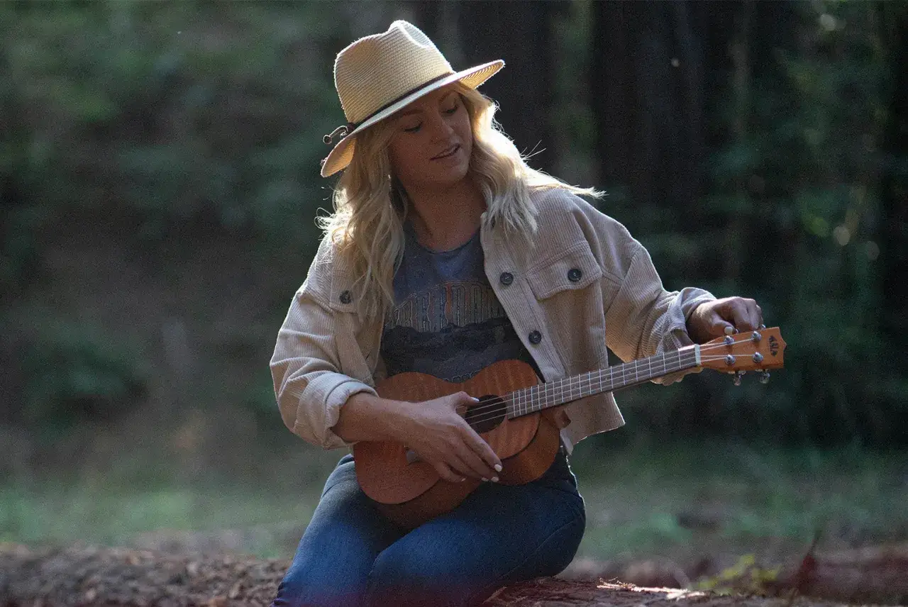 Dziewczyna w kapeluszu nastroiła ukulele sopranowe, siedząc w lesie.