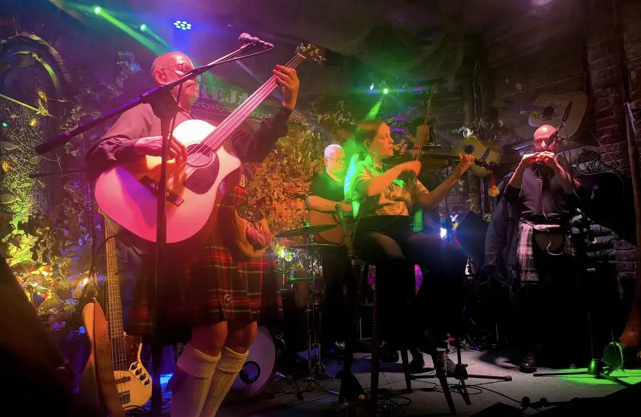 Zespół grający na scenie. W tle wiszą gitary, a muzycy w kiltach wykonują utwory na gitarę i skrzypce.