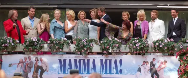 Obsada filmu "Mamma Mia!" pozdrawia fanów. W tle plakat z bohaterami, przypominający o wesołych piosenkach z filmu.