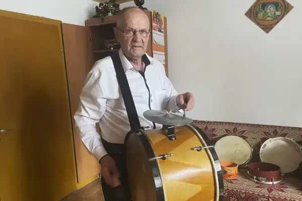 Starszy mężczyzna w białej koszuli gra na dużym, drewnianym bębnie. Obok niego stoją mniejsze instrumenty perkusyjne.