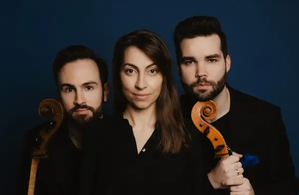 Trzech muzyków, tworzących triolę, pozuje z instrumentami smyczkowymi na tle granatowym.