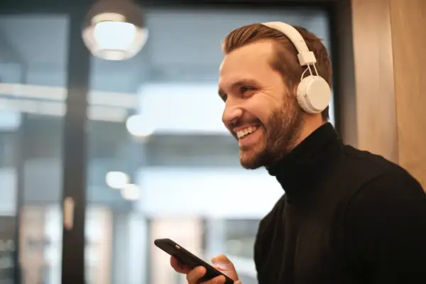 Uśmiechnięty mężczyzna w słuchawkach JBL trzyma telefon, zastanawiając się, jak podłączyć słuchawki JBL do telefonu.